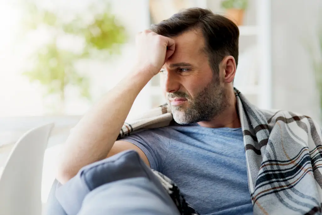 A concerned man sitting on a couch, holding his head in his hands, symbolizing the struggle with mental health. In the background, a supportive figure offers comfort, highlighting the importance of seeking help for mental health check-ups and breaking the stigma surrounding men's mental health.